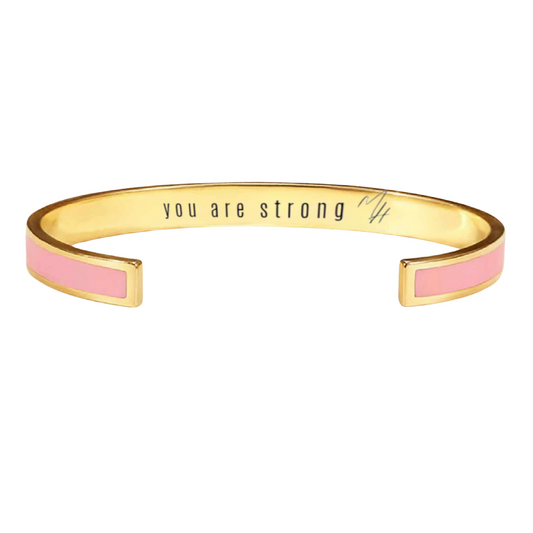 A two-tone gold bangle with pink and white bands, featuring the inscription 'you are strong' on the pink section.